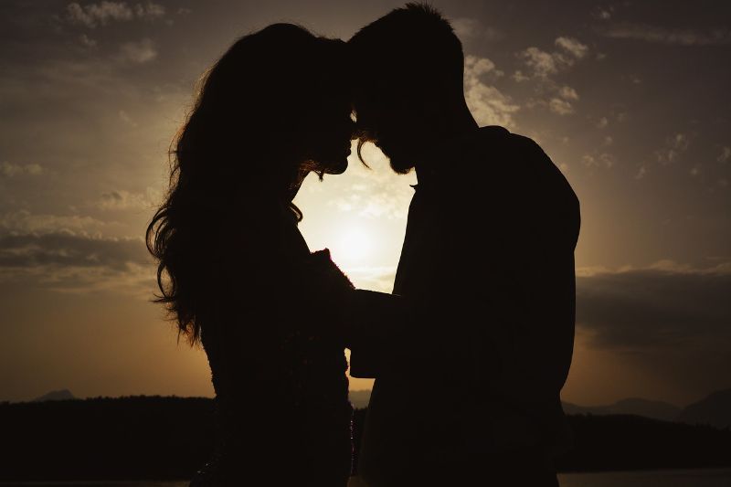 Silhouette einer Frau und eines Mannes vor einem Sonnenuntergang. Sie stehen Stirn an Stirn und halten Händchen.
