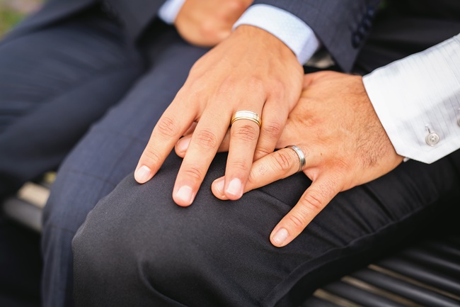 Zwei Männerhände, die auf einem Oberschenkel liegen, beide tragen Verlobungsringe