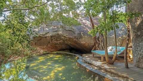 Natürlicher Pool im Schatten großer Felsen und Bäume mit Liegefläche.