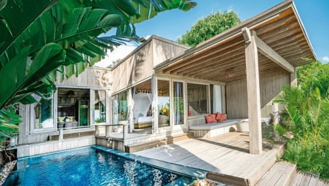 Villa mit Pool, großen Glasfronten und Holzveranda, umgeben von tropischen Pflanzen.