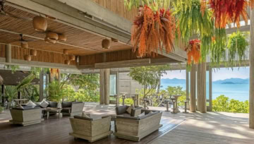 Überdachte Lounge mit Sitzgruppen, Holzdecken und bunten Hängepflanzen, Meerblick im Hintergrund.