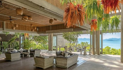 Überdachte Lounge mit Sitzgruppen, Holzdecken und bunten Hängepflanzen, Meerblick im Hintergrund.