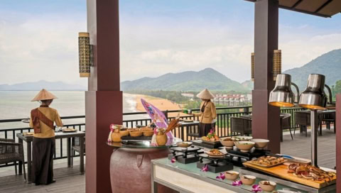 Buffet auf einer Terrasse mit Blick auf Strand und Berge, dekoriert mit Körben und Speisen, zwei Personen in traditioneller Kleidung.