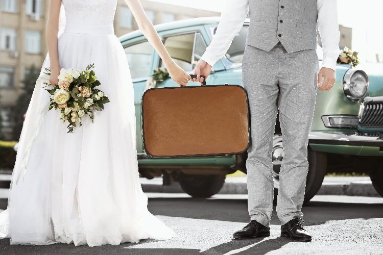 Braut und Bräutigam halten gemeinsam einen Vintage-Koffer in der Hand, bereit für ihre Hochzeitsreise – nostalgische Szene mit Oldtimer im Hintergrund.