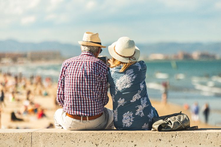 Paar sitzt am Strand mit Blick auf das Meer, sommerliche Urlaubsszene von hinten aufgenommen.