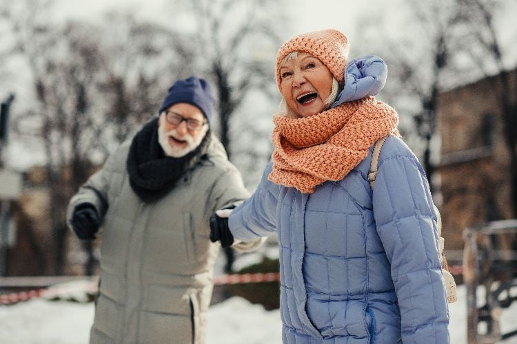 Fröhliches älteres Paar bewegt sich lachend durch eine verschneite Winterlandschaft in warmer Kleidung.