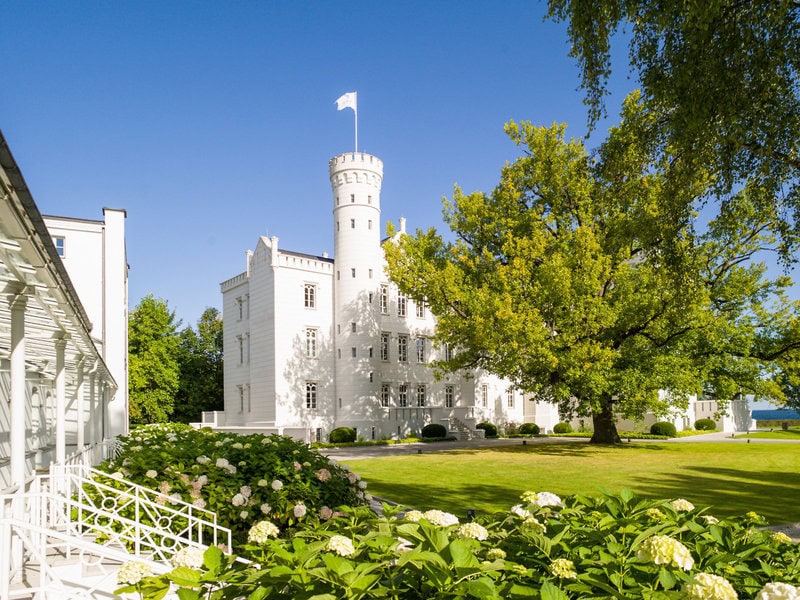 Weißes historisches Gebäude des Grand Hotel Heiligendamm mit Turm, großer Grünanlage, blühenden Hortensien und altem Baumbestand