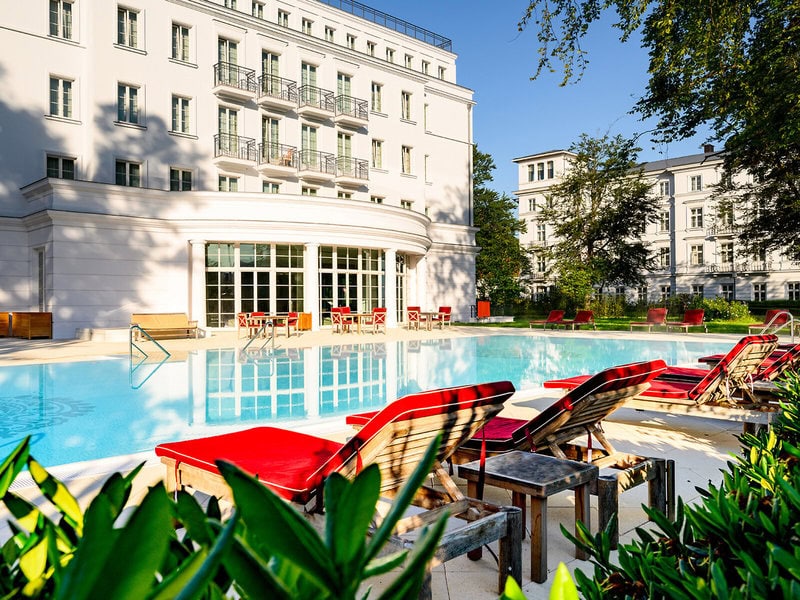 Außenpool des Grand Hotel Heiligendamm mit roten Sonnenliegen, hellen Hotelgebäuden, Spiegelungen im Wasser und grünem Gartenambiente