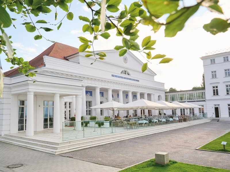 Außenbereich des Restaurants im Grand Hotel Heiligendamm mit weißer Fassade, Säulen, großen Sonnenschirmen und gedeckten Tischen auf der Terrasse