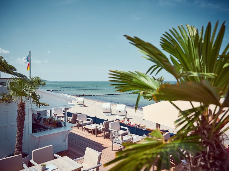 Sonnige Strandterrasse des Grand Hotel Heiligendamm mit Sitzplätzen, Palmen, Strandkörben am Ostseestrand und weitem Blick über das Meer