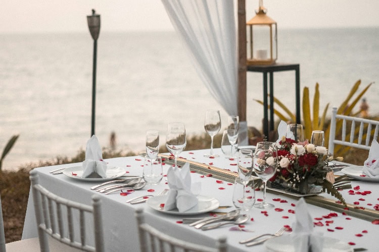 Romantisch gedeckter Tisch am Strand mit Rosenblüten, Gläsern und Blumenarrangement; Meer im Hintergrund.