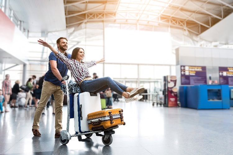 Fröhliches Paar am Flughafen – die Frau sitzt mit ausgebreiteten Armen auf einem Gepäckwagen voller Koffer, während der Mann sie lachend anschiebt.