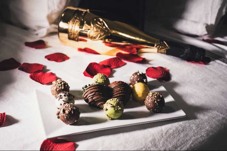 Honeymoon-Setup im Hotel: Pralinen und Schokoherz auf Teller, Rosenblätter auf dem Bett, Champagnerflasche im Hintergrund.