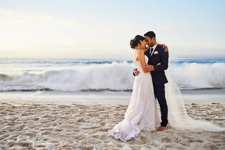 Ein Hochzeitspaar steht am Strand vor einer heranrollenden Welle.