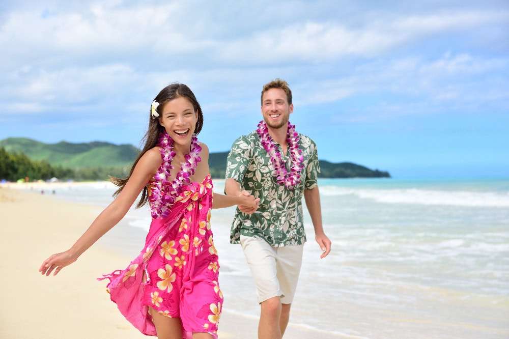Fröhliches Paar mit Blumenketten läuft am Strand von Hawaii entlang.