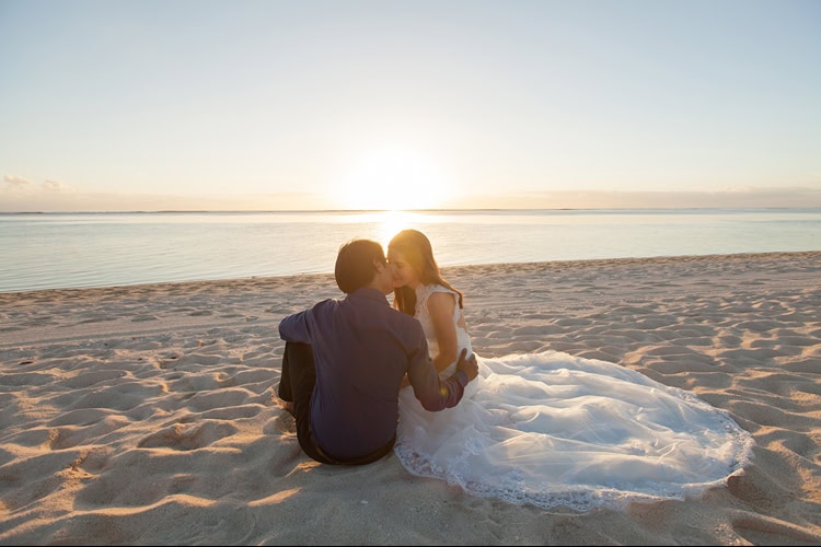 Braut und Bräutigam sitzen bei Sonnenuntergang am Strand und küssen sich, das Brautkleid liegt im Sand neben dem Paar.