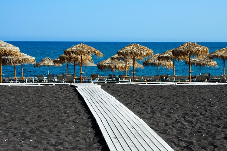 Schwarzer Vulkanstrand mit Strohliegen und Blick auf das tiefblaue Meer – typisch für Santorini.