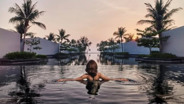 Frau schwimmt im Infinity-Pool bei Sonnenuntergang mit Palmen im Hintergrund