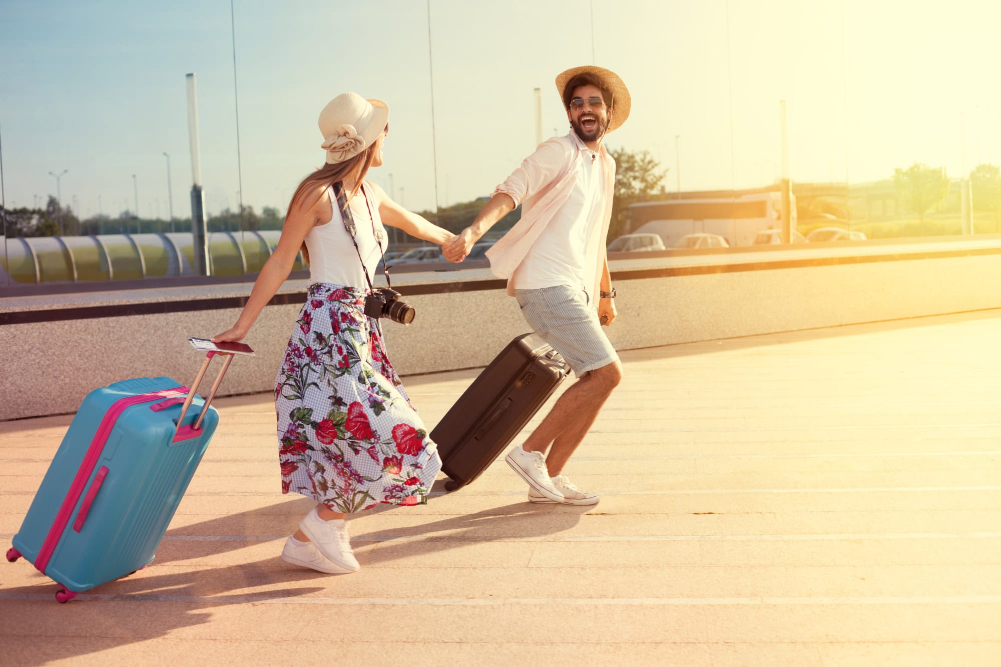 Fröhliches Paar mit Koffern auf dem Weg in den Urlaub, sie halten sich an den Händen und lachen, während sie durch die Sonne laufen – Kamera und Reiselust im Gepäck.