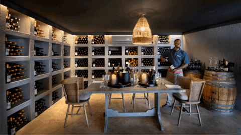 Weinraum mit gefüllten Regalen, Verkostungstisch und Gastgeber beim Einschenken