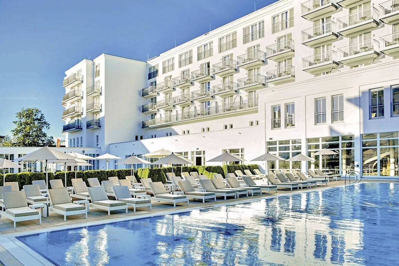 Außenpool des Steigenberger Grandhotel & Spa Heringsdorf mit weißen Sonnenliegen, Sonnenschirmen, heller Hotelfassade und spiegelndem Wasser im Vordergrund