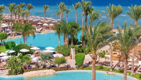 Poolanlage des Hotels aus der Vogelperspektive. Man sieht die Palmen und das Meer. Dazwischen die vielen Sonnenschirme und Poolliegen.